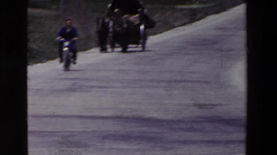 A man on a motorcycle approaches along a pale rural road while a cart or carriage and other figures appear in the distance, consistent with archival home-movie footage from Spain in 1969.