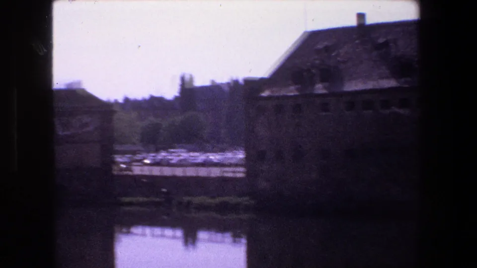 1970 Strasbourg 8mm frame with old buildings at the edge of still water, including a large weathered structure and distant parked cars or light-toned vehicles.