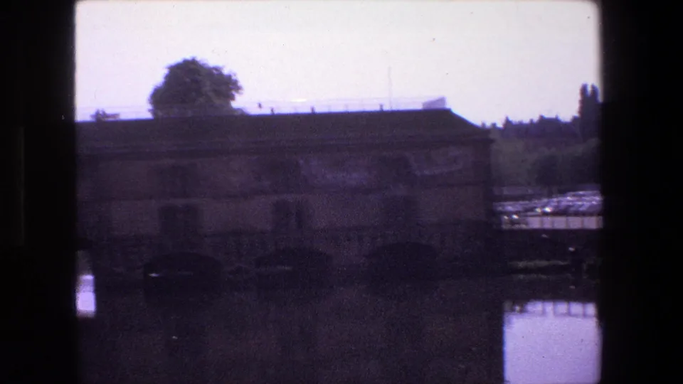 1970 Strasbourg 8mm view of a low weathered waterside building reflected in muddy still water, with roofline details and trees beyond.