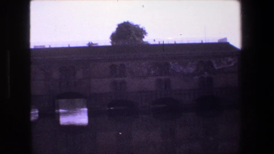 1970s 8mm view in Strasbourg showing a stone bridge with arched openings over dark water, with a tree and low skyline beyond.
