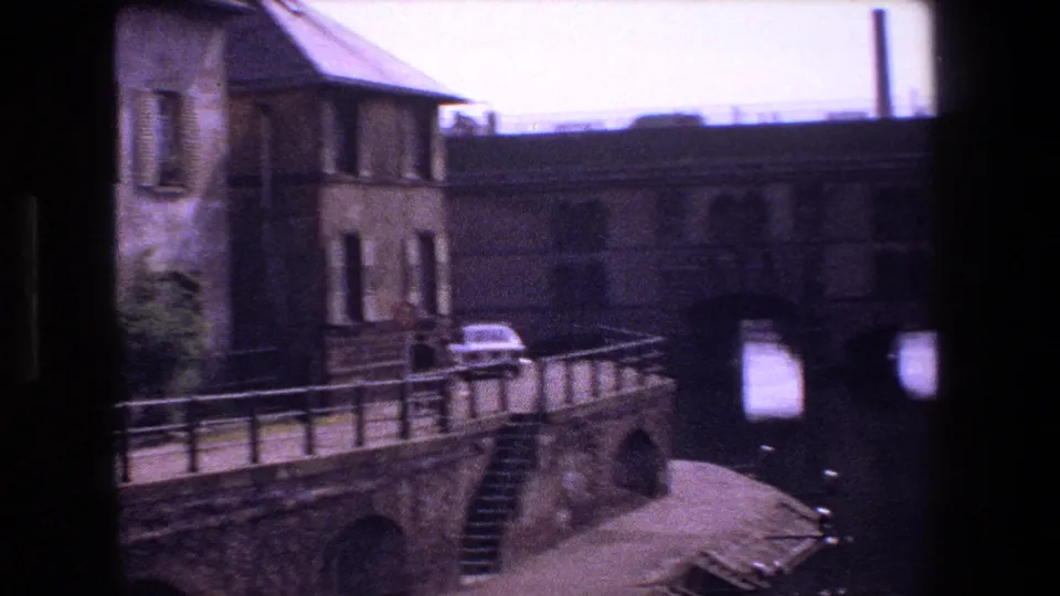 1970s Strasbourg riverside view with a masonry building, bridge railing, white car, steps down to the water, and dark bridge arches in the background.
