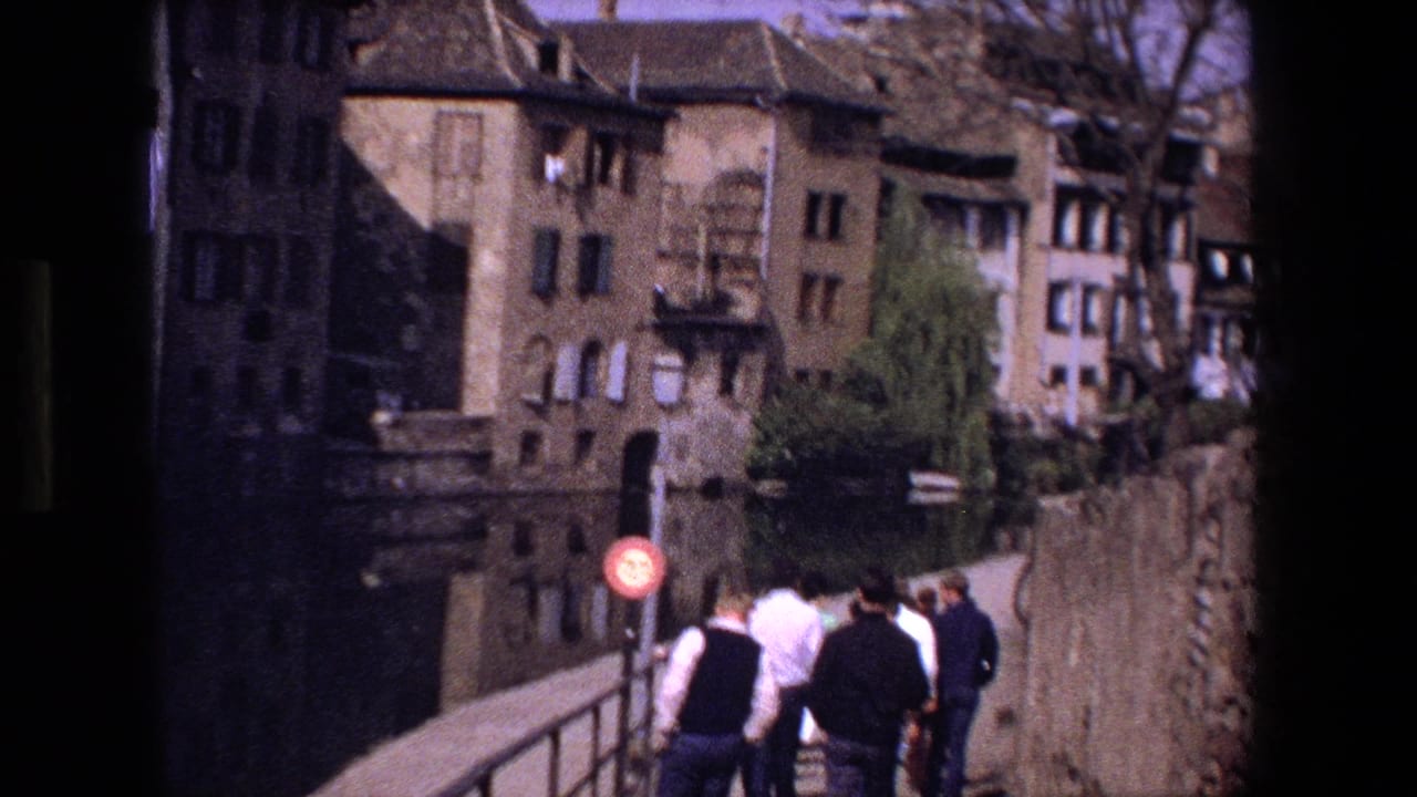 In Strasbourg, France, 1970 home-movie footage, several pedestrians walk along a stone bridge with shuttered historic buildings, bare trees, and canal-side walls behind them.