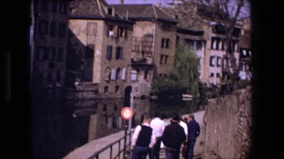 In Strasbourg, France, 1970 home-movie footage, several pedestrians walk along a stone bridge with shuttered historic buildings, bare trees, and canal-side walls behind them.