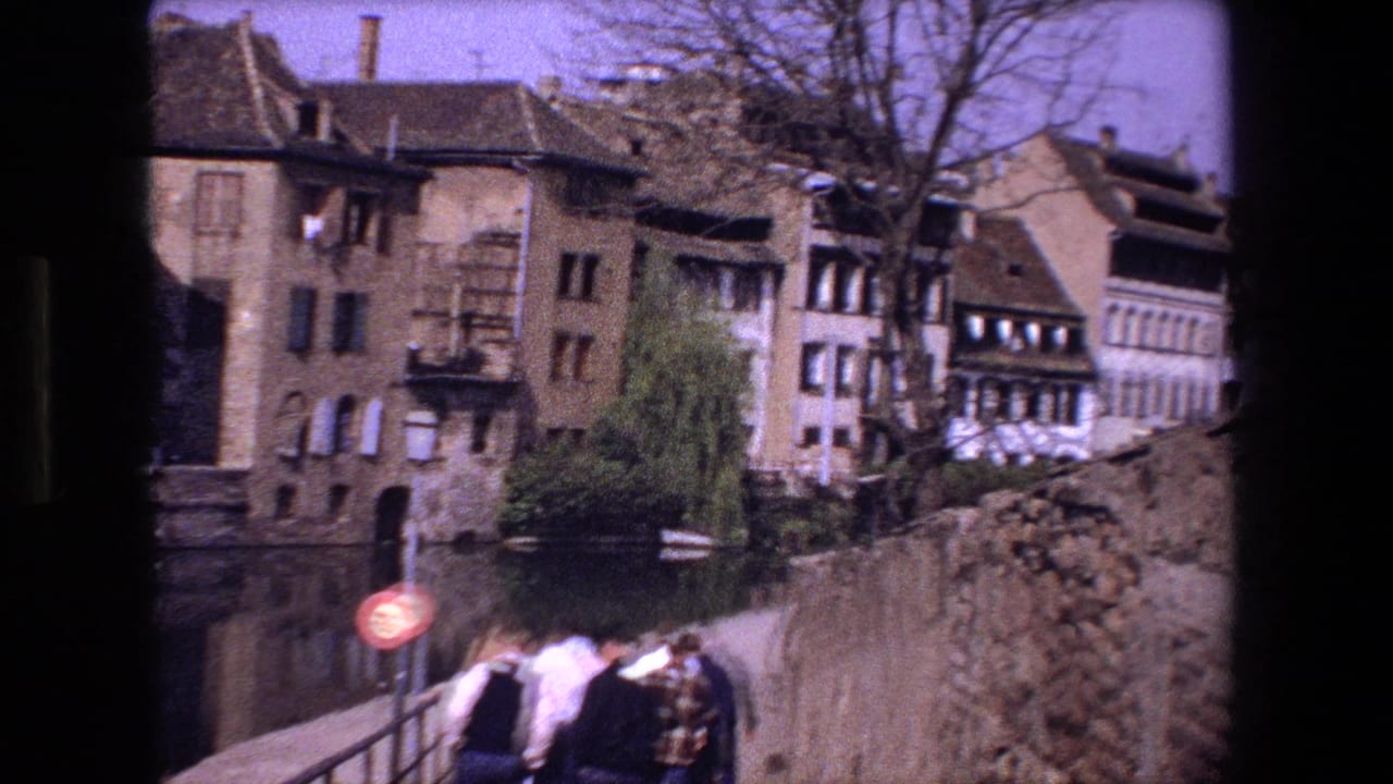 A group walks along the bridge with old buildings and a stone wall in the background.
