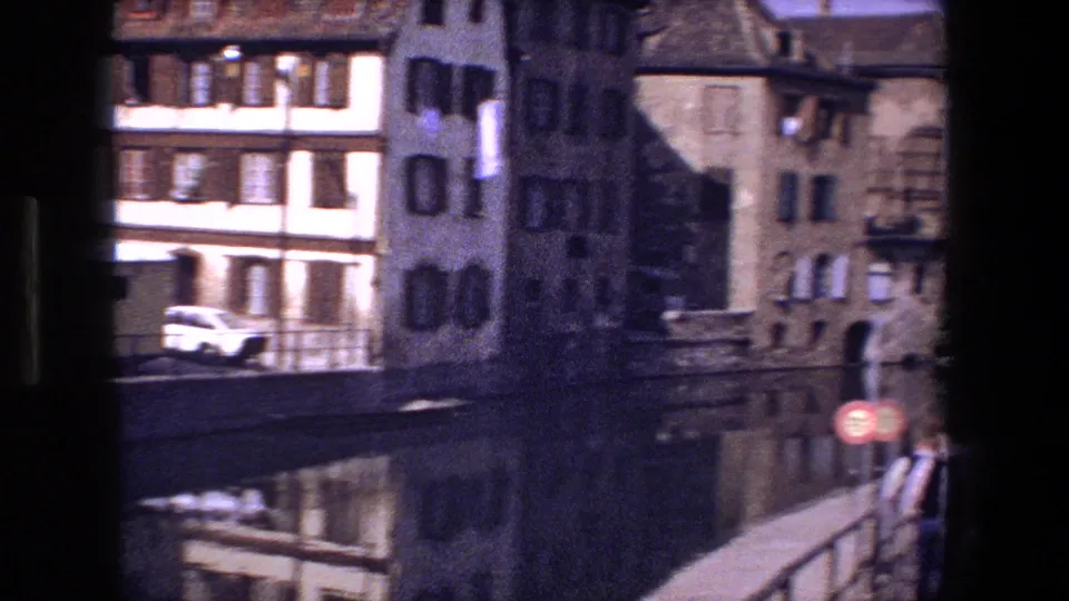 The frame shows canal reflections and old Strasbourg buildings, with a white compact car, bridge railings, and a red circular traffic sign visible near the water.
