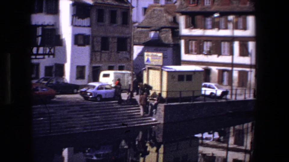 A 1970 Strasbourg street view shows stone steps, parked period cars, pedestrians, a small yellow kiosk, and timber-framed and shuttered buildings near the canal.