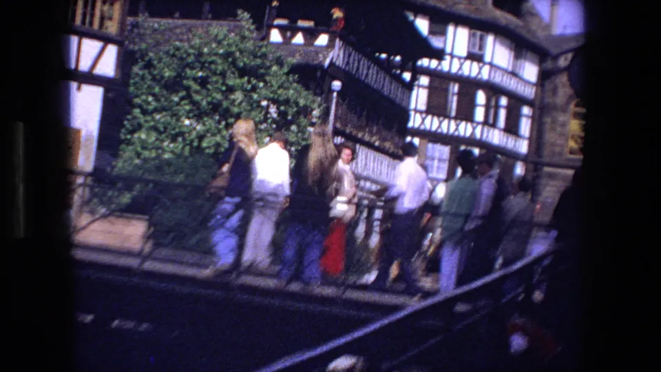 1970 8mm frame from Strasbourg showing visitors on a bridge near half-timbered buildings, with railings and leafy greenery in bright daylight.
