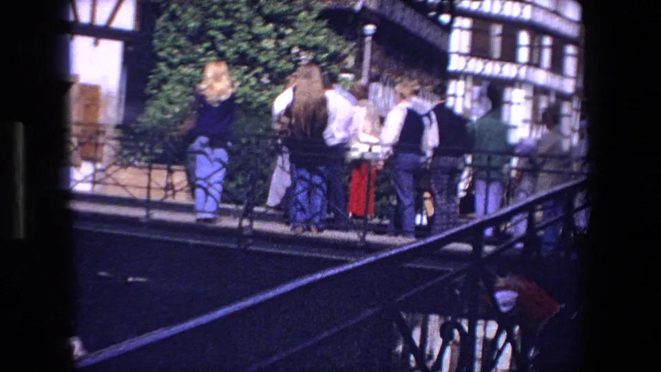 1970 Strasbourg home-movie frame showing several visitors on a bridge beside half-timbered facades and a leafy tree or shrub.