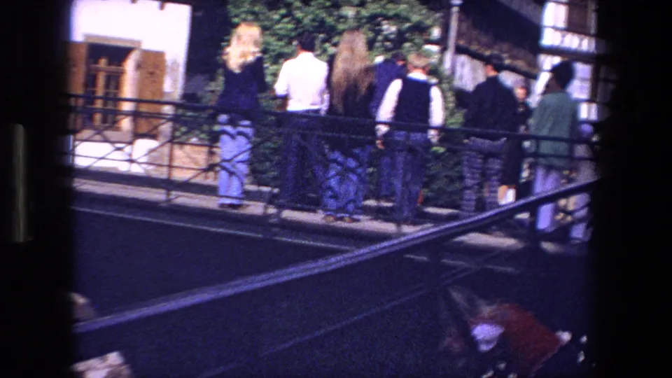1970 8mm frame showing visitors from behind on a Strasbourg bridge, with metal railings, a leafy wall, and traditional half-timbered building details nearby.
