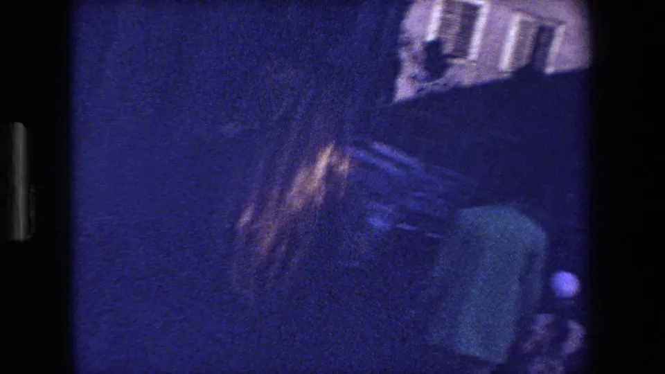 Grainy 1970 8mm Strasbourg night-street frame with a blurred pedestrian near a building facade and dark roadway.