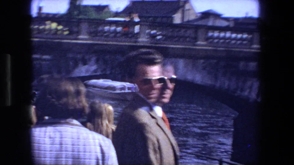 1970 Strasbourg sightseeing moment with tourists near canal water, including a man in a jacket and sunglasses turning toward the handheld camera.