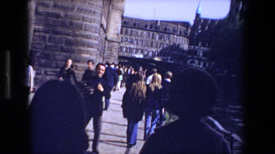 1970 Strasbourg canal walkway with pedestrians in coats moving past stone architecture, bridge shadows, and city buildings in a handheld home-movie frame.