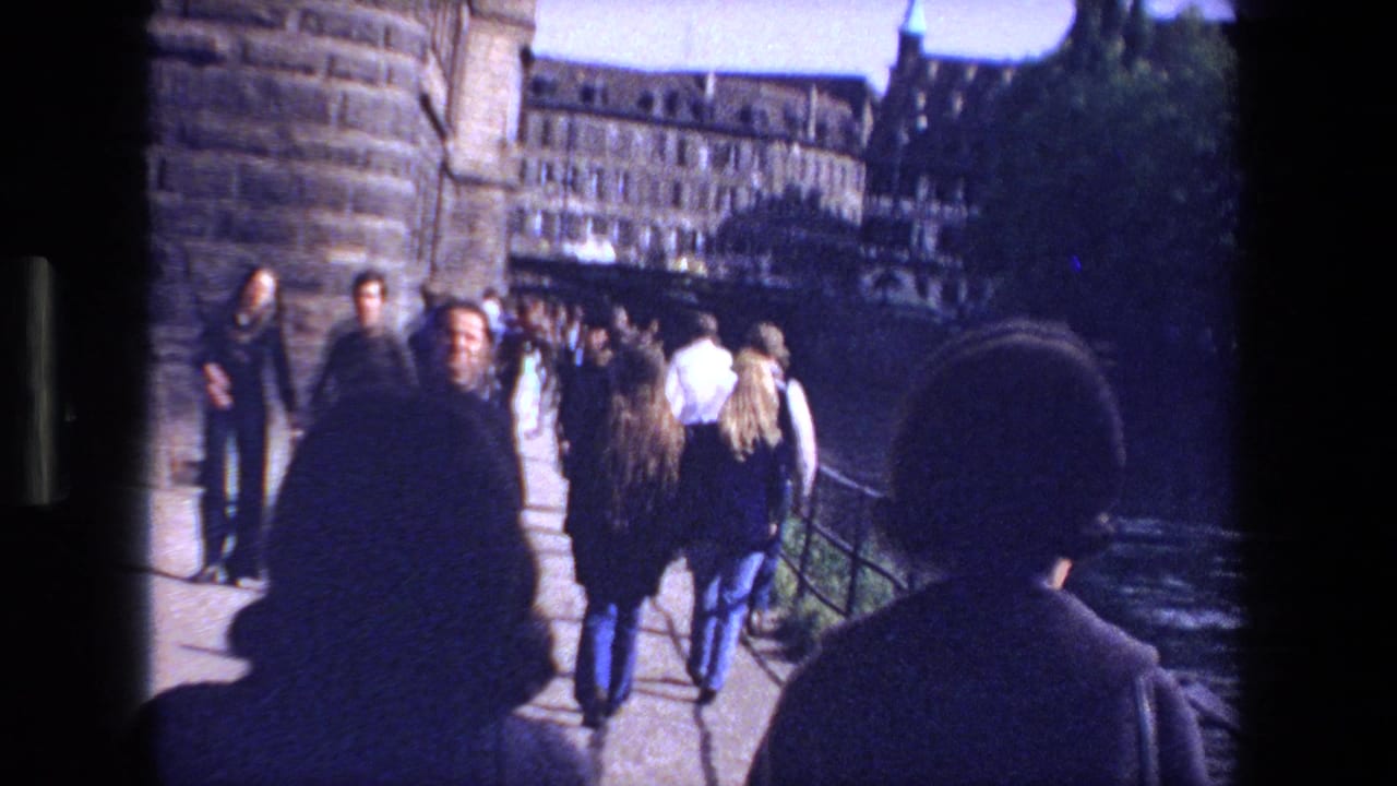 Pedestrians walk across a stone bridge beside a river with older buildings in the background.