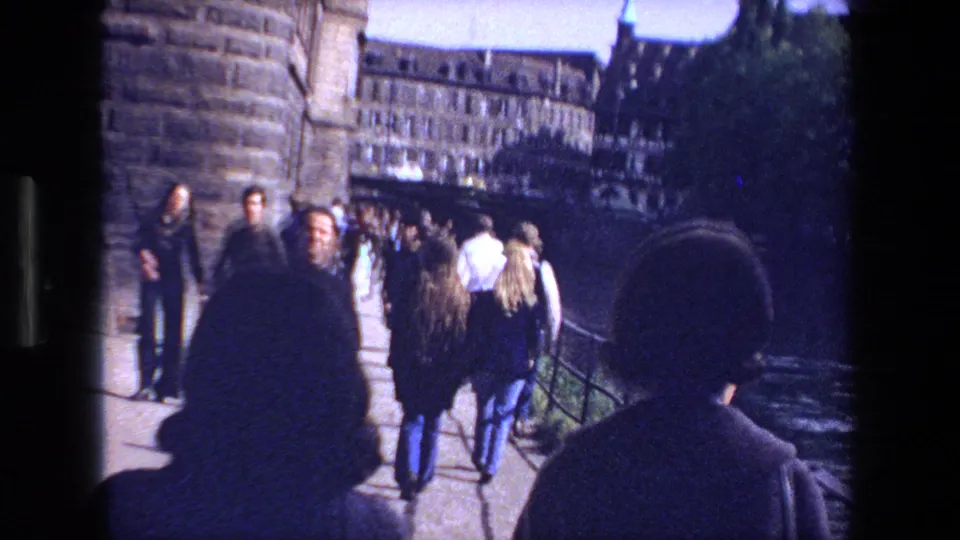 In Strasbourg, France, pedestrians in 1970s-era clothing walk along a stone bridge beside the river, with older city buildings beyond them.