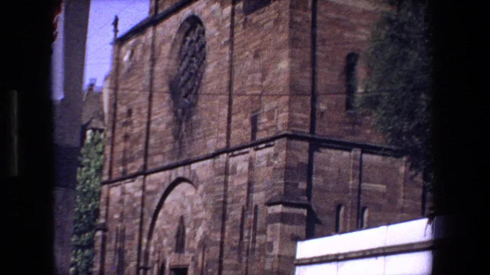 1970 8mm frame showing the lower stone church exterior in Strasbourg with an arched doorway, rose window, white barrier, and nearby street details.