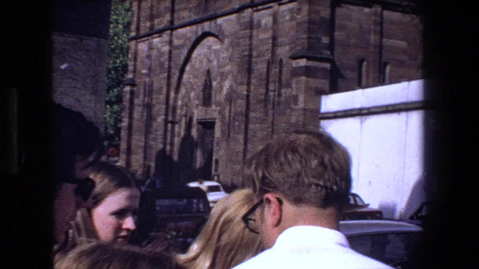 1970 Strasbourg street scene with tourists in the foreground, parked cars, and a historic stone church facade behind them.