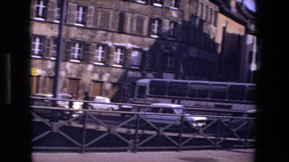 1970 Strasbourg street frame with a bridge railing in the foreground, cars and a bus passing, stone buildings behind, and a distant pedestrian on the sidewalk.