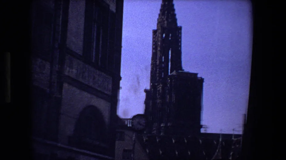1970 home-movie view in Strasbourg, France, with a Gothic cathedral spire, clock face, and shadowed architectural facade filling the frame.