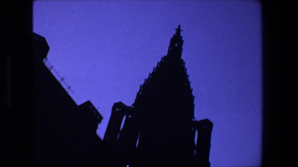 1970s 8mm Strasbourg, France frame showing a tall Gothic cathedral spire and roofline in silhouette against a deep blue twilight sky.
