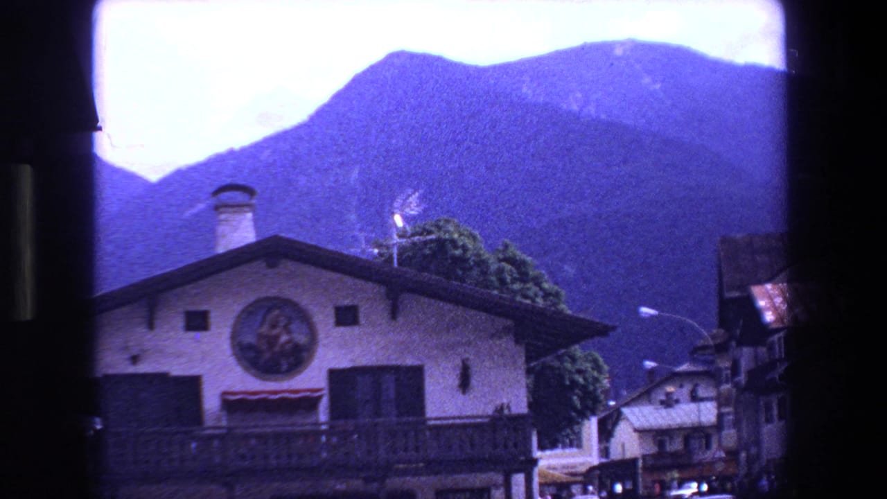 Building facade with circular painting, dark shutters, balcony, and mountains behind it.