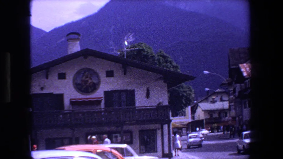 A 1970 Strasbourg street view with a painted building facade, compact cars, pedestrians, shopfronts, and mountains visible beyond the rooftops.