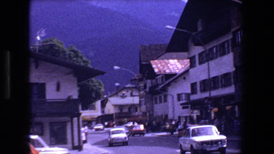 A wider 1970 Strasbourg street frame showing buildings along both sides, several compact cars, pedestrians near the sidewalks, and mountains behind the town.