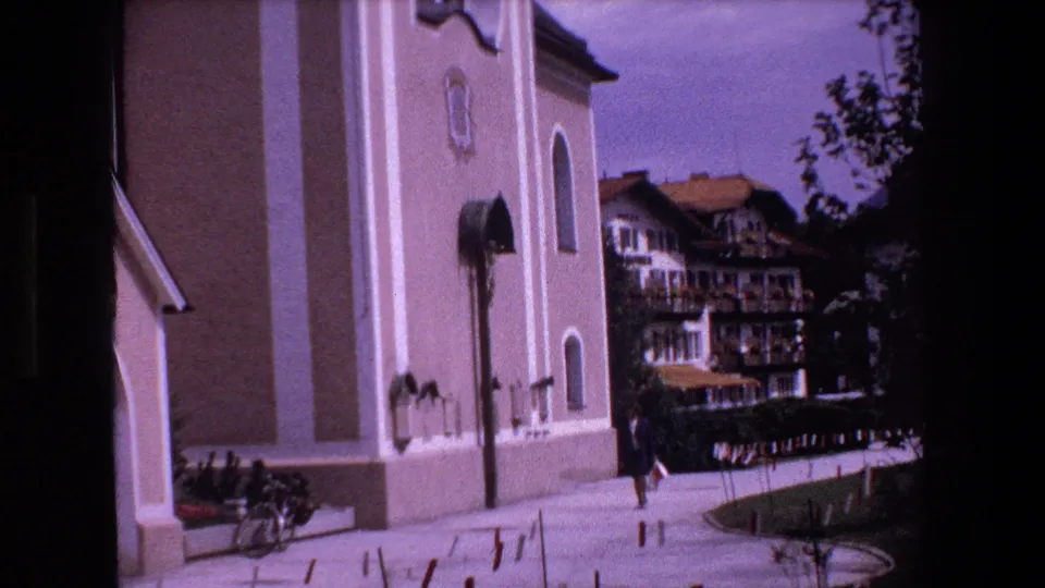 1970 Strasbourg home-movie frame showing a pale church exterior, arched windows, a pedestrian on the walkway, bicycles, and multi-story buildings beyond.
