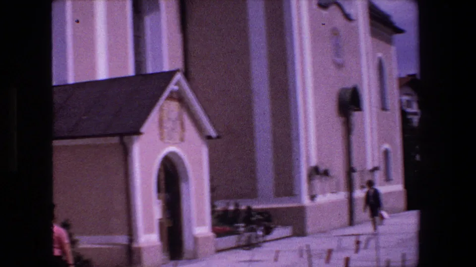 1970 Strasbourg home-movie frame showing the church entrance, pale facade, bicycles near the wall, and a pedestrian moving through the foreground.