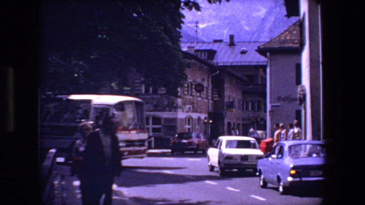 Street view with a bus, parked cars, pedestrians, and town buildings.