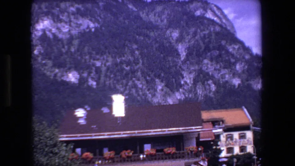 Metadata-listed French valley-town view with a roadside building, balcony flowers, dark roof, trees, and steep wooded mountains behind it.