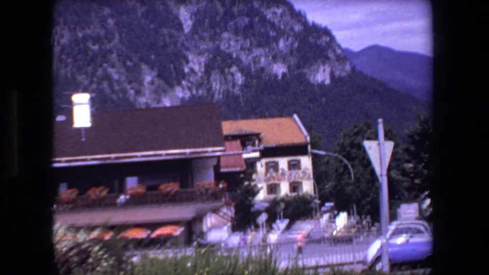 1970 8mm roadside view in the metadata-listed French location, with chalet-like buildings, a triangular road sign, a car, fencing, and wooded mountains in the background.