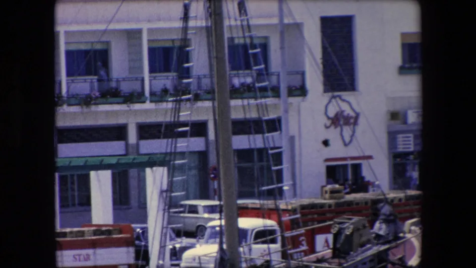 1971 Algeria home-movie frame showing sailboat mast rigging in the foreground, a white multi-story building with balconies, an Africa outline graphic, and cars parked below.