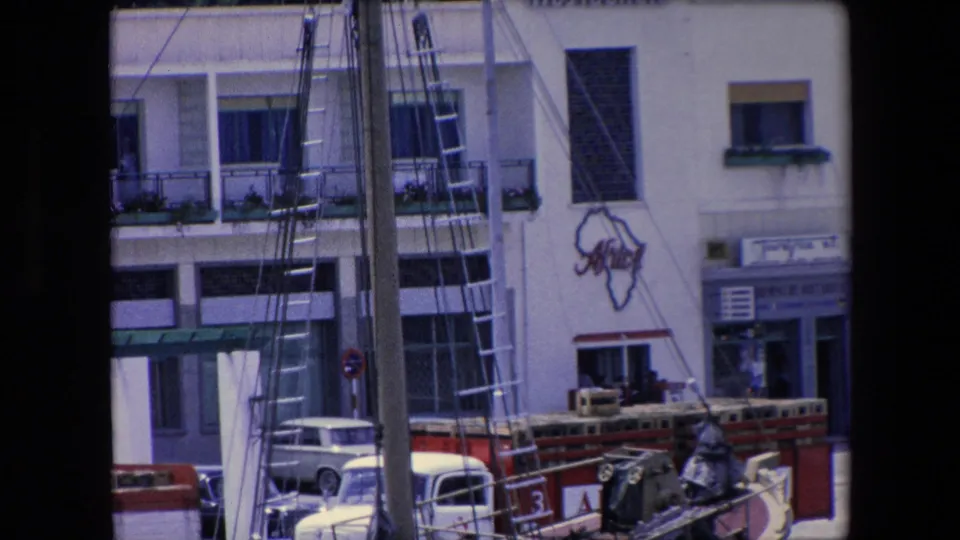 1971 Algeria frame from a harbor viewpoint with mast rigging in front of a white building, balcony planters, parked cars, and a painted Africa-shaped graphic on the wall.