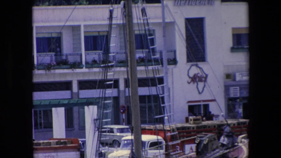 1971 Algeria home-movie view with sailboat rigging crossing a white building facade, balconies with flower boxes, shuttered windows, and parked 1970s cars below.