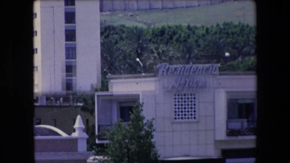 1971 Algeria 8mm frame showing a distant white waterfront building with cursive signage, geometric wall openings, nearby apartment windows, and greenery behind dark frame edges.