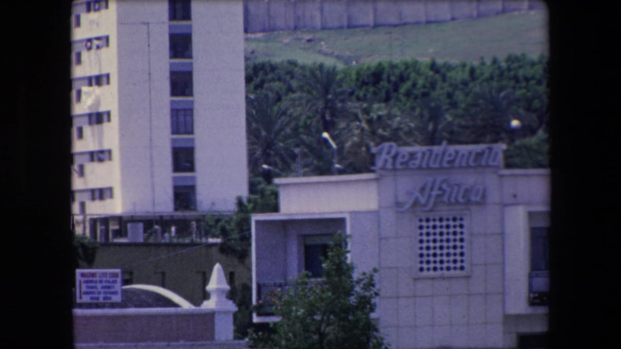 1971 Algeria 8mm frame with a white modernist waterfront building, cursive lettering on the facade, a walled foreground, trees, and a partial tower behind it.