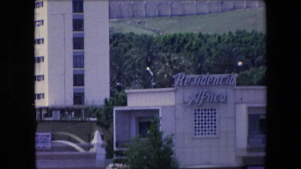 1971 Algeria 8mm frame showing the same waterfront building from a slightly lower view, with cursive signage, lattice-like wall detail, greenery, and a tower edge.