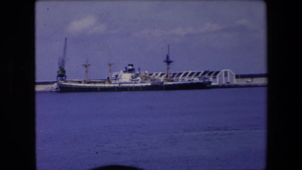 1971 Algeria 8mm frame showing a docked vessel at a pier across calm blue water, with low harbor structures and a cloudy sky in the background.