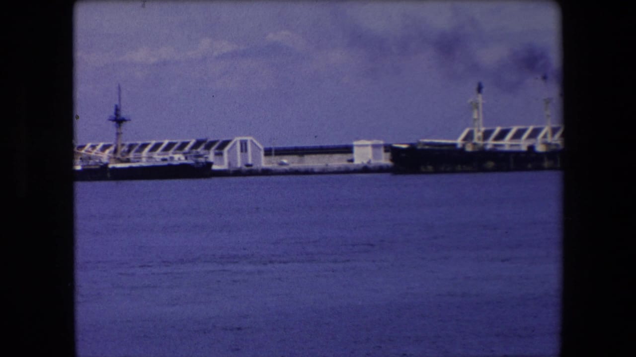 Distant view of a large ship and dock structures across blue harbor water in archival 1971 Algeria home-movie footage.