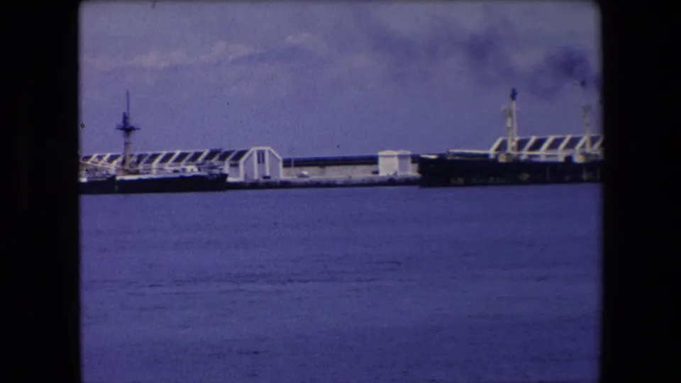 Distant view of a large ship and dock structures across blue harbor water in archival 1971 Algeria home-movie footage.