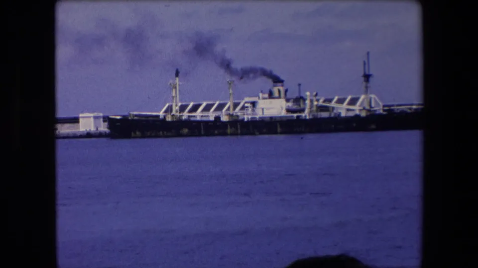 Large dark-hulled ship moored by a pier in Algeria, with black smoke rising from the funnel over calm blue water.