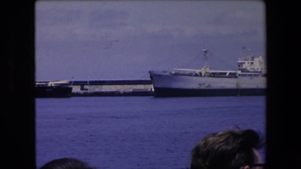 A white ship or forward section sits beside the pier, with partial spectators' heads visible along the lower foreground of the home-movie frame.