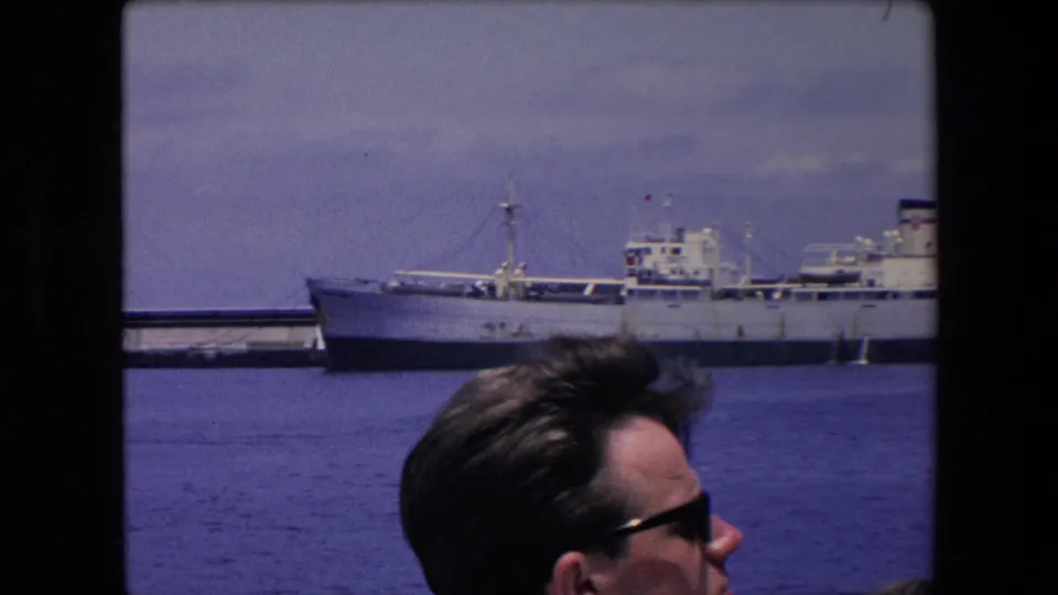 1971 Algeria home-movie frame showing an adult man in sunglasses partly visible beside a dark window frame, with a large cargo vessel docked across blue harbor water.