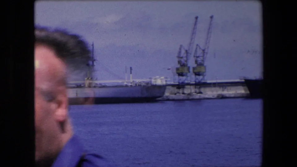 1971 Algeria home-movie frame with a blurred adult face close to the camera, harbor water beyond, dock cranes, and part of a cargo vessel along the pier.