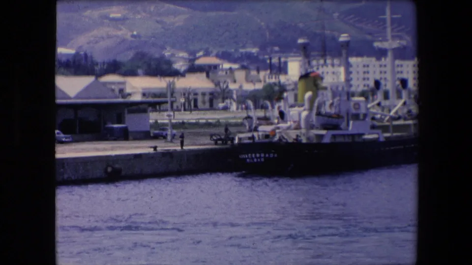 1971 Algeria home-movie frame showing a harbor quay, a dark-hulled vessel with small visible lettering, industrial waterfront buildings, and hills rising behind the port.