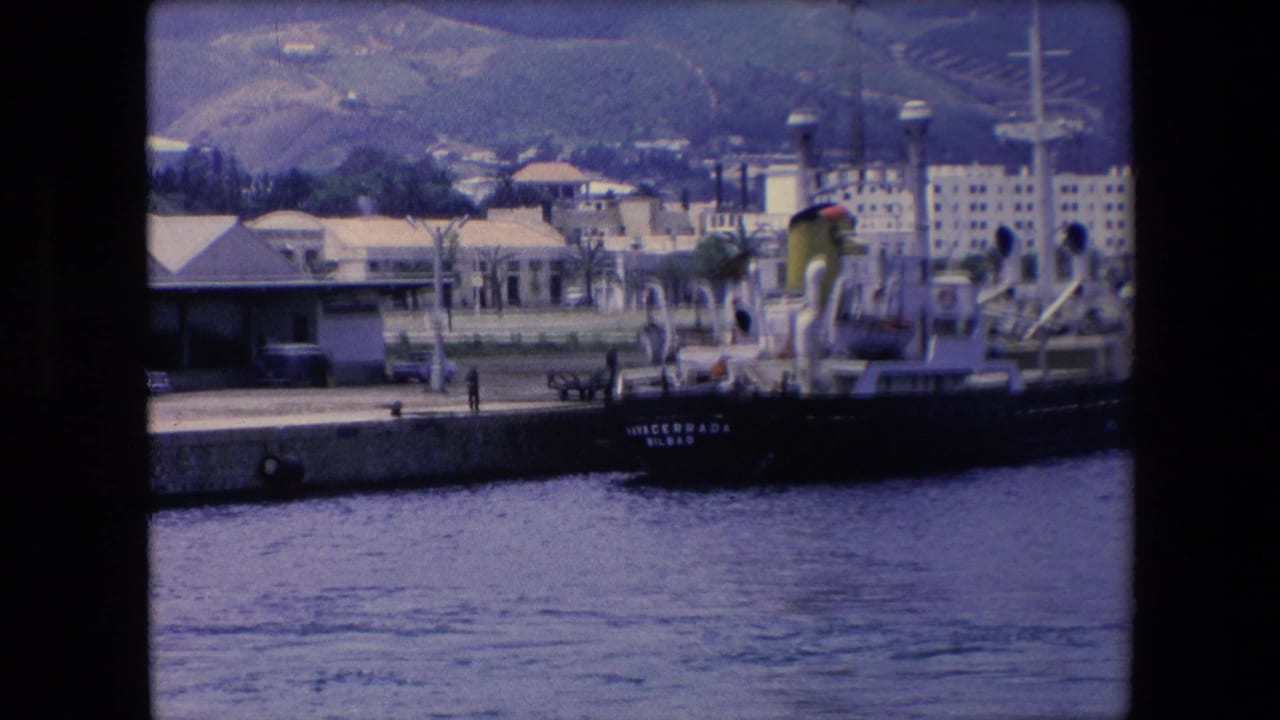 1971 8mm travel frame showing a dark-hulled cargo ship at a concrete pier, with waterfront buildings, palm trees, water, and hills in the background.