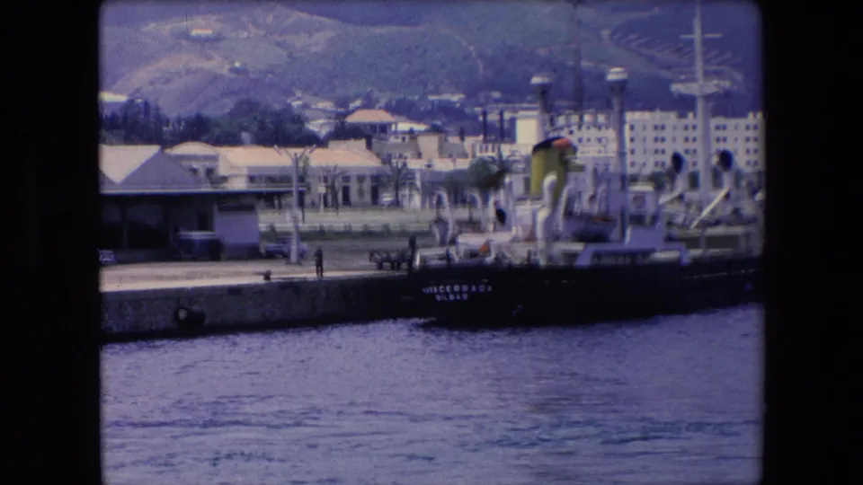1971 8mm travel frame showing a dark-hulled cargo ship at a concrete pier, with waterfront buildings, palm trees, water, and hills in the background.