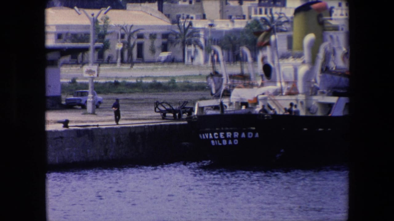 Closer 8mm frame of the NAVACERRADA at the pier, with a lone pedestrian, a small car, palm trees, and waterfront buildings visible behind the ship.