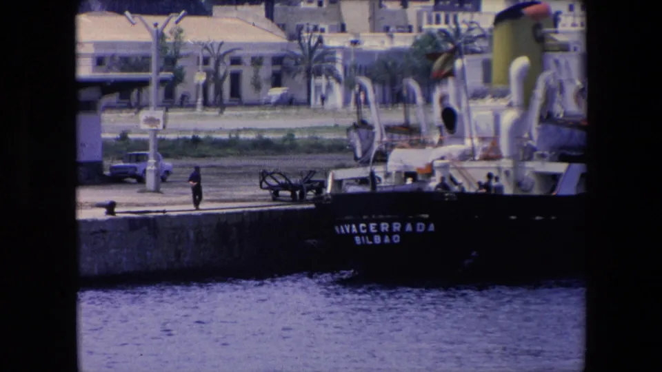 Closer 8mm frame of the NAVACERRADA at the pier, with a lone pedestrian, a small car, palm trees, and waterfront buildings visible behind the ship.