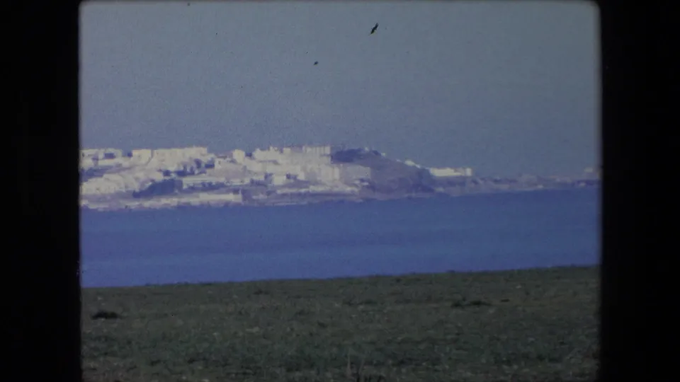 Distant coastal view from the same 8mm travel sequence, showing a white hillside town across blue water with grass in the foreground and haze over the shoreline.